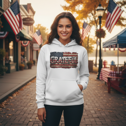 Woman wearing a white hoodie with 'GRATEFUL' and American flag design, standing on a street with American flags in the background.