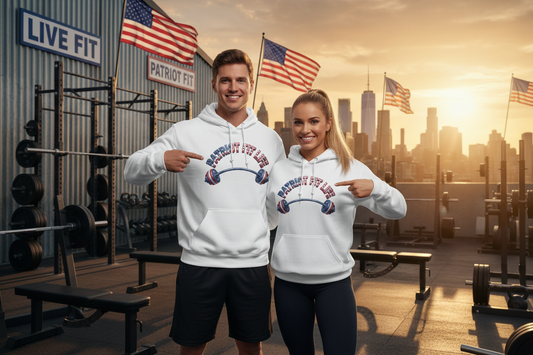 A man and a woman standing on a rooftop gym set at sunset, wearing matching white hoodies with a patriotic graphic that reads 'Patriot Fit Life' with an American flag and barbell design. The background features weight racks, weight benches, and multiple American flags against a city skyline.