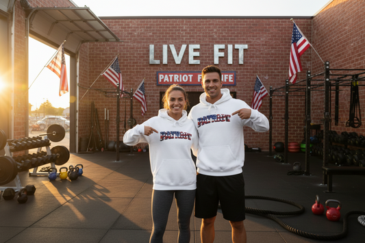 A man and a woman are standing in an outdoor gym wearing matching white hoodies with 'PATRIOT' printed on the front, and they are both pointing at the logos. The background shows a brick wall with 'LIVE FIT' & PATRIOT FIT LIFE signage, American flags, and gym equipment.