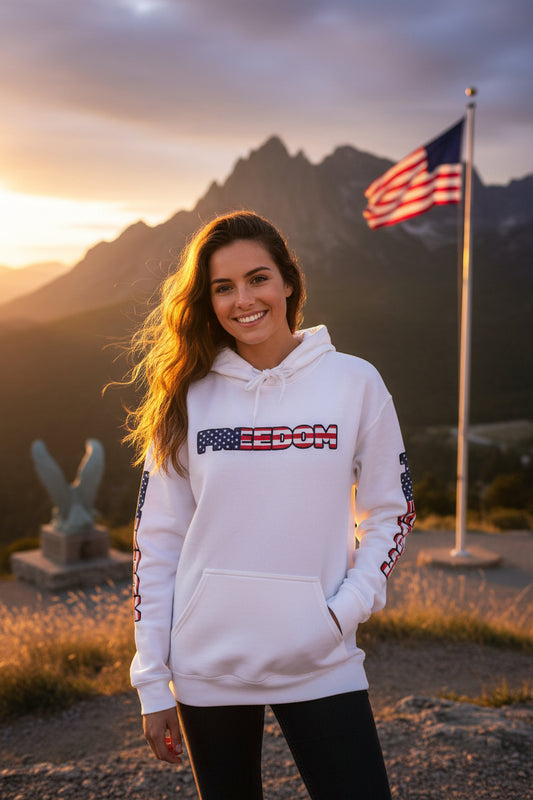 A woman wearing a white hoodie with 'FREEDOM' printed on it, standing against an American flag and mountainous landscape during sunset.