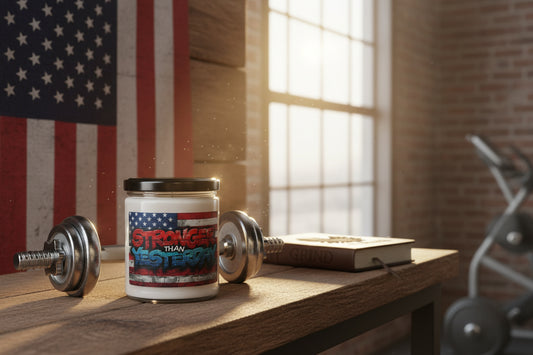 Candle with the Stronger Than Yesterday American flag design on a wooden surface with dumbbells and a notebook.
