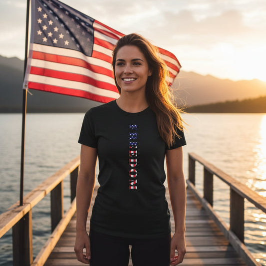 Woman wearing a black t-shirt with a vertical flag design, standing on a dock by a lake with an American flag in the background.