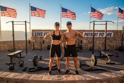 A shirtless man and a woman stand together on an outdoor fitness platform with American flags, weight machines, kettlebells, battle ropes, and a stone wall behind them. The woman wears a black sports bra and fitted shorts, and the man wears black shorts and a black cap.