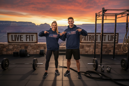 Two people standing on a rooftop gym at sunset wearing matching navy blue hoodies with a patriotic red, white, and blue embroidered PATRIOT graphic across the chest. Barbell racks, battle ropes, and stone signs reading 'LIVE FIT' and 'PATRIOT FIT LIFE' are visible against the Grand Canyon.