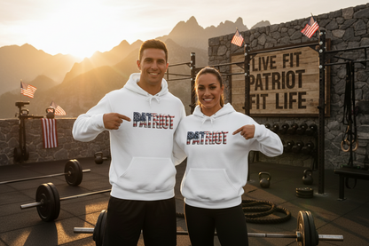 A man and a woman stand in a rustic mountain gym wearing matching white hoodies with a patriotic 'PATRIOT' graphic in red, white, and blue. The background features a stone wall, a wooden sign reading 'LIVE FIT PATRIOT FIT LIFE', and barbell racks under a warm sunset sky.