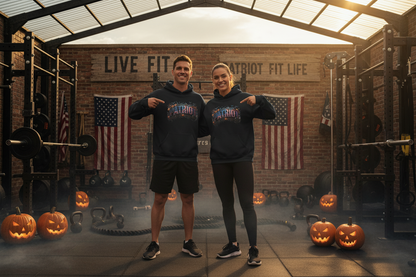 Two people standing in a gym wearing matching black hoodies with a Halloween PATRIOT graphic across the chest, pointing to the logos. The background features a brick wall with American flags, weight racks, kettlebells, and jack-o'-lanterns.