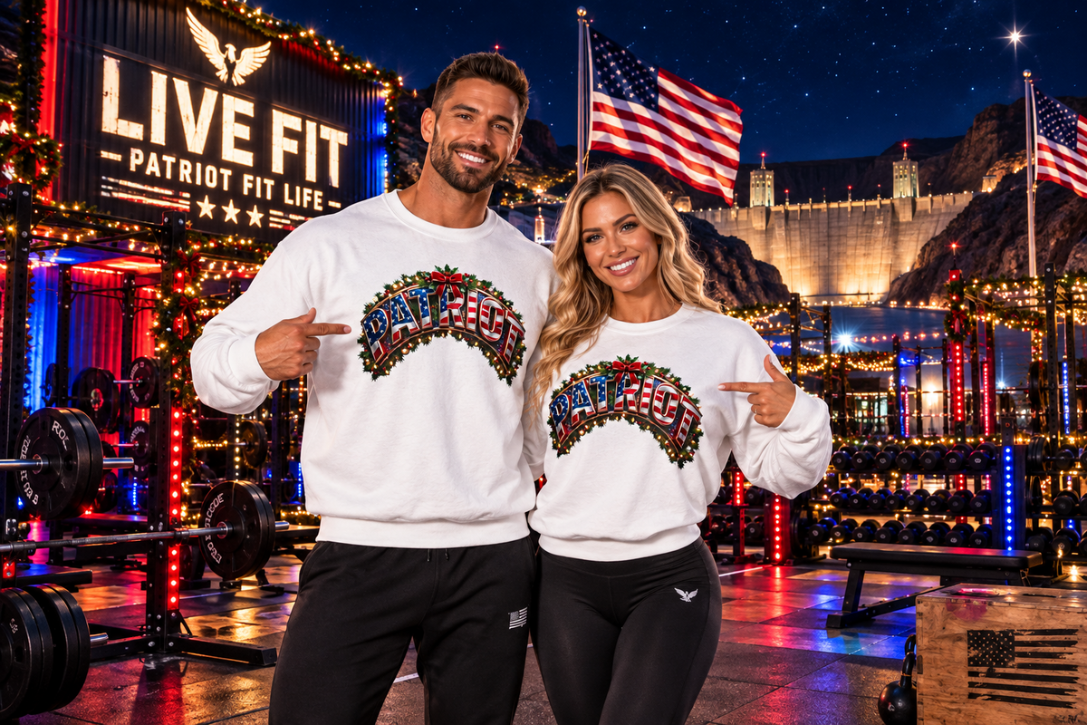 A man and a woman stand in a brightly lit gym at night wearing matching white crewneck sweatshirts with a colorful 'PATRIOT' graphic across the chest. Behind them is the Hoover Dam, American flags, a large 'LIVE FIT PATRIOT FIT LIFE' sign, weight equipment, and mountains under a starry sky.
