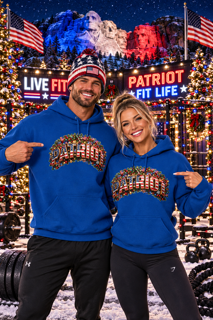 A man and a woman posing together in a snowy, festive outdoor gym setting, both wearing matching royal blue hoodies with a large festive 'PATRIOT' graphic across the chest. The background features Mount Rushmore illuminated in red, white, and blue at night, with American flags, Christmas lights, and 'LIVE FIT' and 'PATRIOT FIT LIFE' signs.