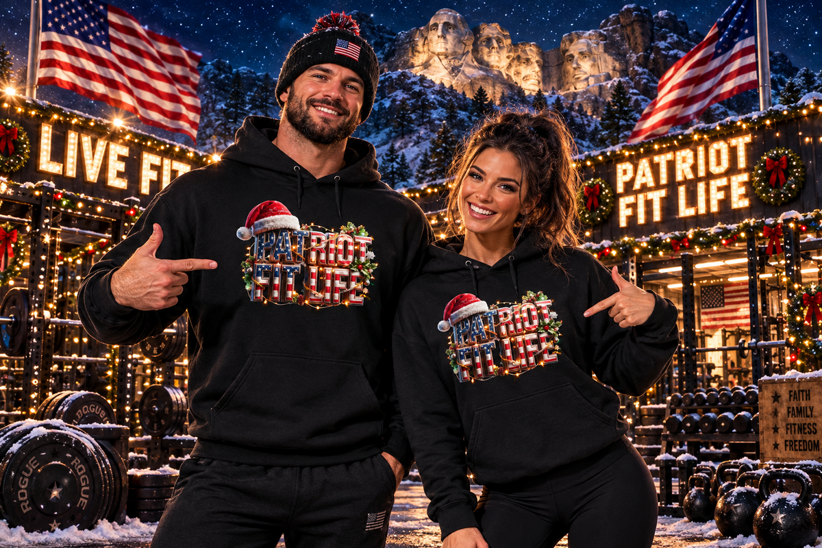 A man and a woman wearing matching black hoodies with a patriotic Christmas graphic that reads PATRIOT FIT LIFE, standing in front of a festive outdoor gym at night. The background features snow-covered Mount Rushmore, American flags, and lit signs reading 'LIVE FIT' and 'PATRIOT FIT LIFE.'