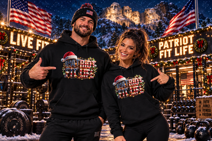A man and a woman wearing matching black hoodies with a patriotic Christmas graphic that reads PATRIOT FIT LIFE, standing in front of a festive outdoor gym at night. The background features snow-covered Mount Rushmore, American flags, and lit signs reading 'LIVE FIT' and 'PATRIOT FIT LIFE.'