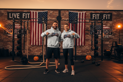 A man and a woman standing in a gym wearing matching light gray hoodies with a 'PATRIOT' Halloween design on the front, posing in front of American flags and gym equipment.