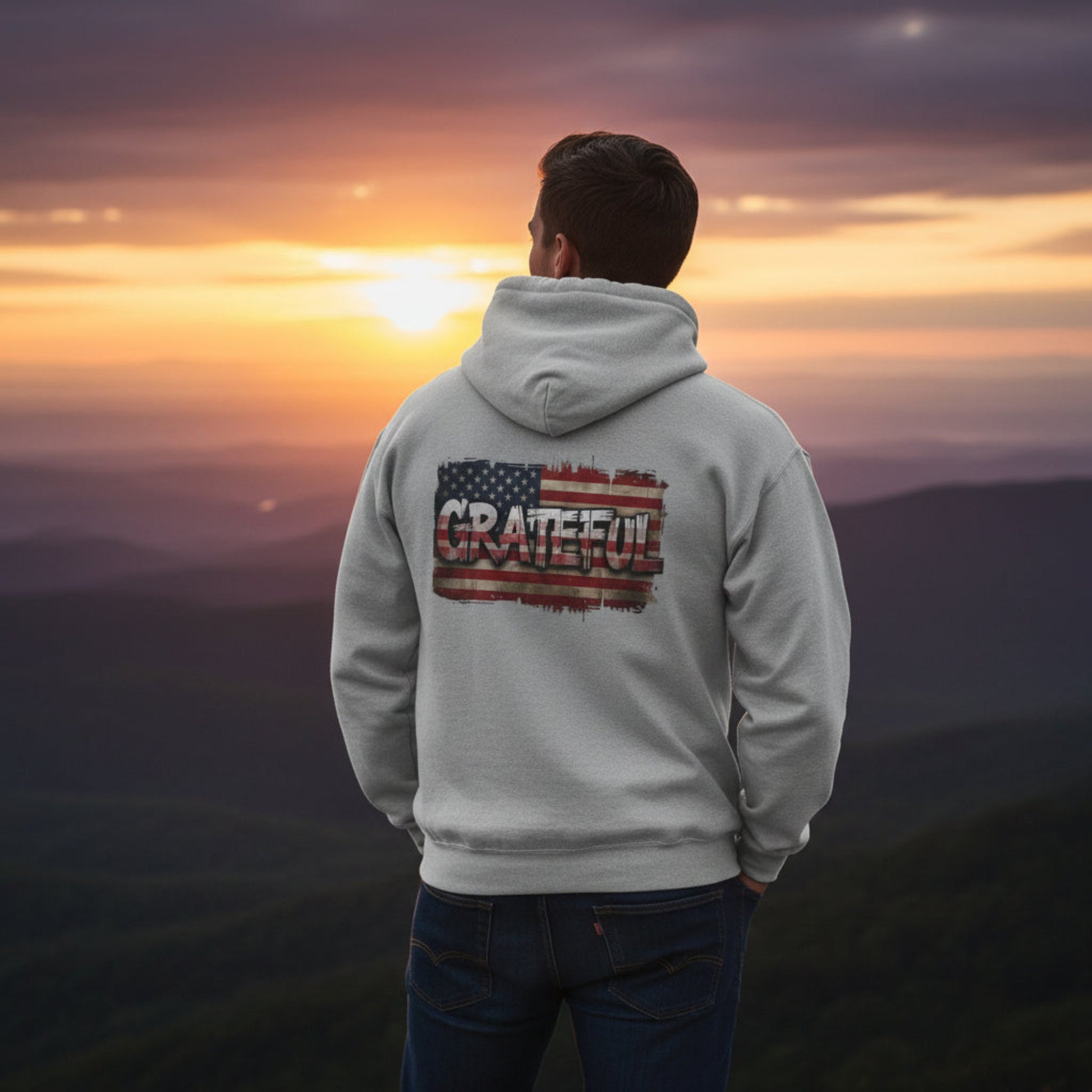 Person wearing a hoodie with 'Grateful' and an American flag design, standing against a sunset landscape.
