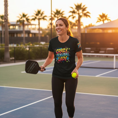 A woman wearing a black t-shirt with colorful 'Crazy Pickleball Lady' text and graphics standing on a pickleball court.