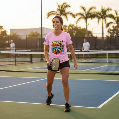 A woman on a pickleball court wearing a pink 'Crazy Pickleball Lady' shirt with palm trees and mountains in the background.