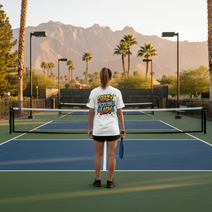 A woman standing on a pickleball court wearing a t-shirt that says Crazy Pickleball Lady with mountains and palm trees in the background.