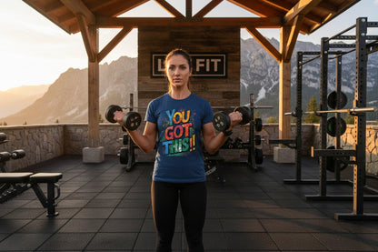 A woman exercising with dumbbells wearing  a blue t-shirt with You Got This!! text in a mountainous outdoor setting.