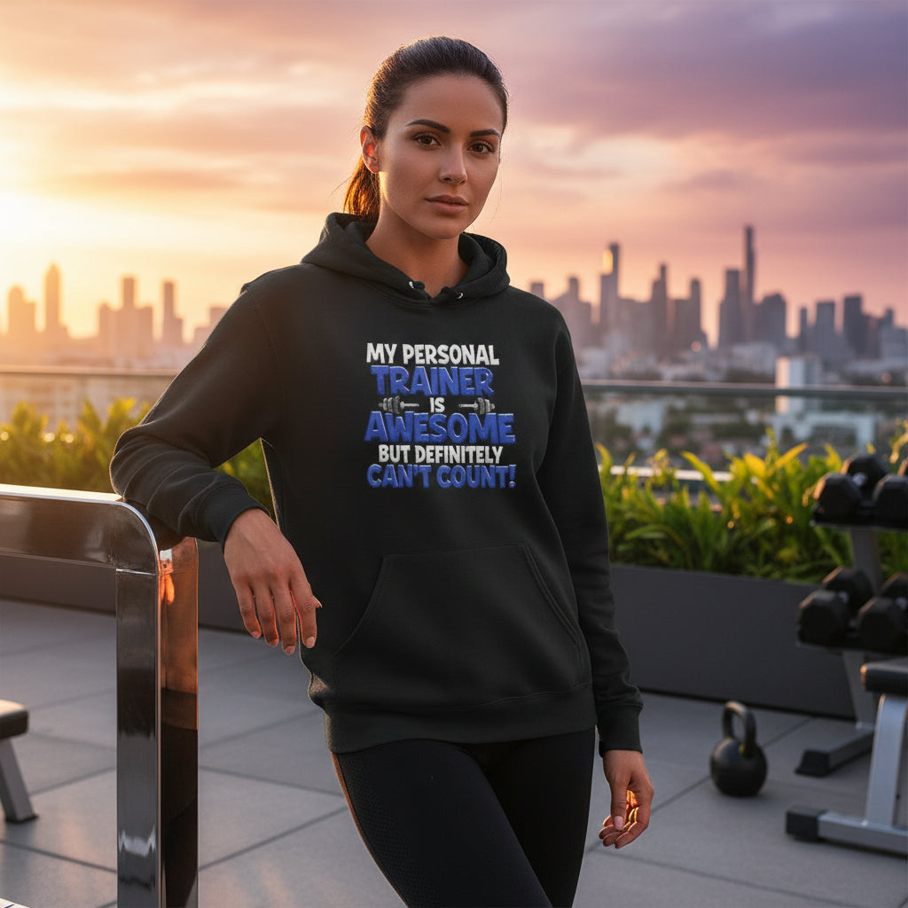 A woman wearing a black hoodie with text on it that says, My Personal Trainer Is Awesome But Definitely Can't Count!, standing in an outdoor gym with a city skyline in the background.