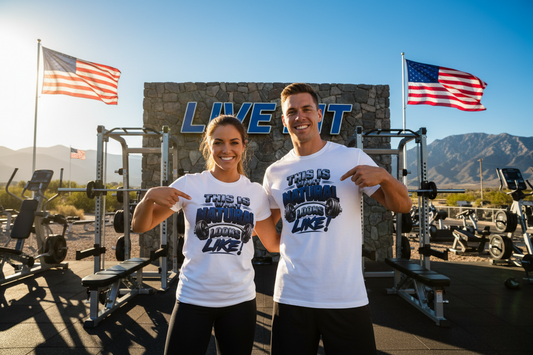 Two smiling people are standing in a gym in front of a stone wall with the words “LIVE FIT,” wearing matching white graphic T-shirts that read “THIS IS WHAT NATURAL LOOKS LIKE!” with a barbell graphic. American flags, exercise equipment, mountains, and a blue sky are visible in the background.