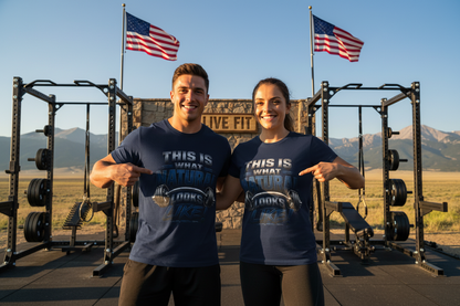 Two smiling people standing outdoors in front of a rustic wooden sign that reads 'LIVE FIT' with mountains and open land in the background. Both are wearing matching navy graphic T-shirts with white and light blue text that says 'THIS IS WHAT NATURAL LOOKS LIKE,' pointing at the shirts.