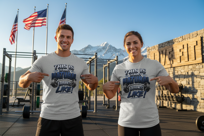 Two smiling people are standing in a gym outdoors, wearing matching light gray T-shirts with a bold graphic that reads 'THIS IS WHAT NATURAL LOOKS LIKE! in dark blue and black lettering. Flags, mountains, and gym equipment are visible in the background.