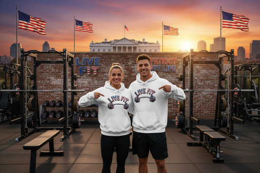 A male and a female are standing in an outside gym wearing matching white hoodies with a 'LIVE FIT' American flag lettering pattern over an American flag lettering pattern barbell graphic on the front; they both are pointing at the designs on the front of their hoodies. The background features a brick wall with 'LIVE FIT & PATRIOT FIT LIFE' lettering, signs, weight racks, and American flags against the White House and a sunset sky.