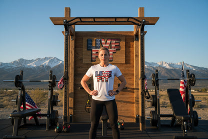 A woman wearing a t-shirt that says LIVE FIT standing in front of a wooden fitness structure with mountains in the background.
