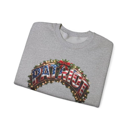 A folded heather gray crewneck sweatshirt featuring a festive graphic on the front. The graphic displays the word 'PATRIOT' in arched, illuminated marquee-style letters filled with an American flag pattern, framed by a green holiday wreath with red berries and warm string lights, all on a clean white background.