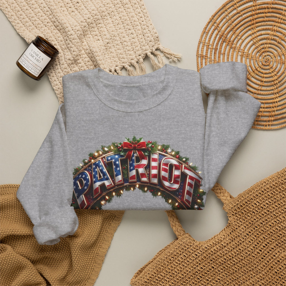 A gray crewneck sweatshirt laid flat on a light beige surface, featuring a graphic print of the word 'PATRIOT' in red, white, and blue American flag lettering with holiday garland and lights.
