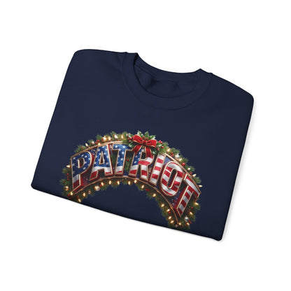 A navy blue crewneck sweatshirt folded flat, featuring a festive graphic on the front with the word 'PATRIOT' in 3D letters decorated with an American flag pattern, surrounded by green pine branches, red berries, and warm glowing Christmas lights, set against a plain white background.