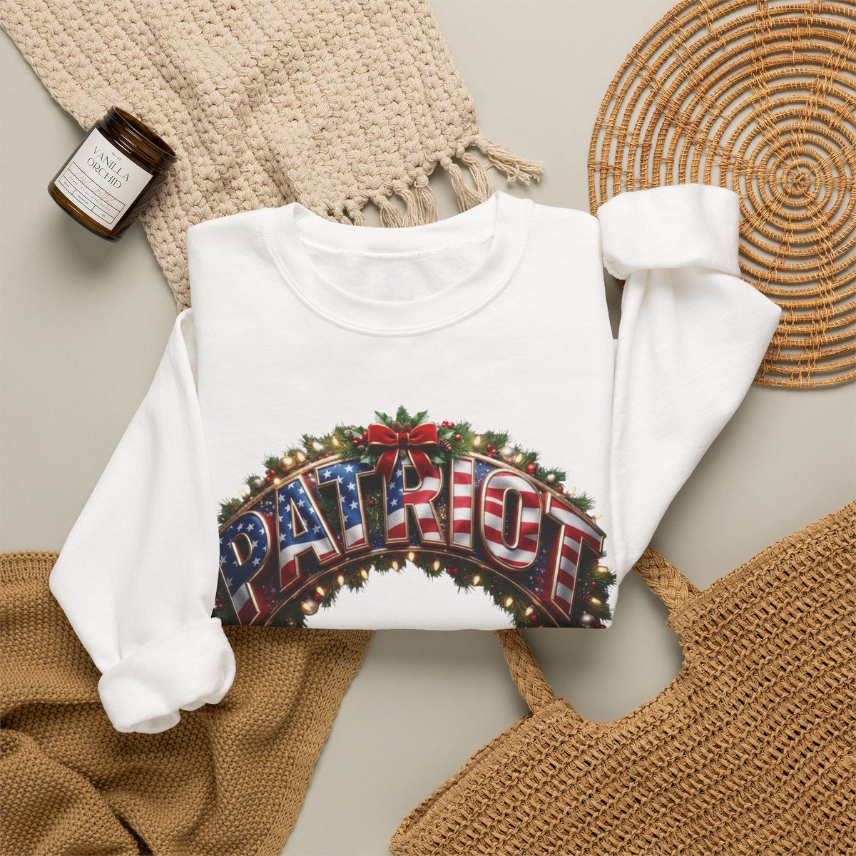 A white long-sleeve crewneck sweatshirt with a festive graphic reading 'PATRIOT' in red, white, and blue flag lettering under a green holiday wreath.