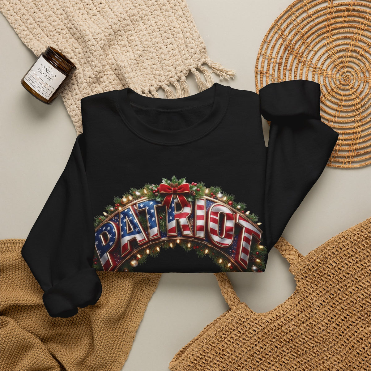 A black long-sleeve crewneck sweatshirt laid flat on a light beige surface, featuring a large graphic print that reads 'PATRIOT' in an American flag pattern with holiday lights and greenery.