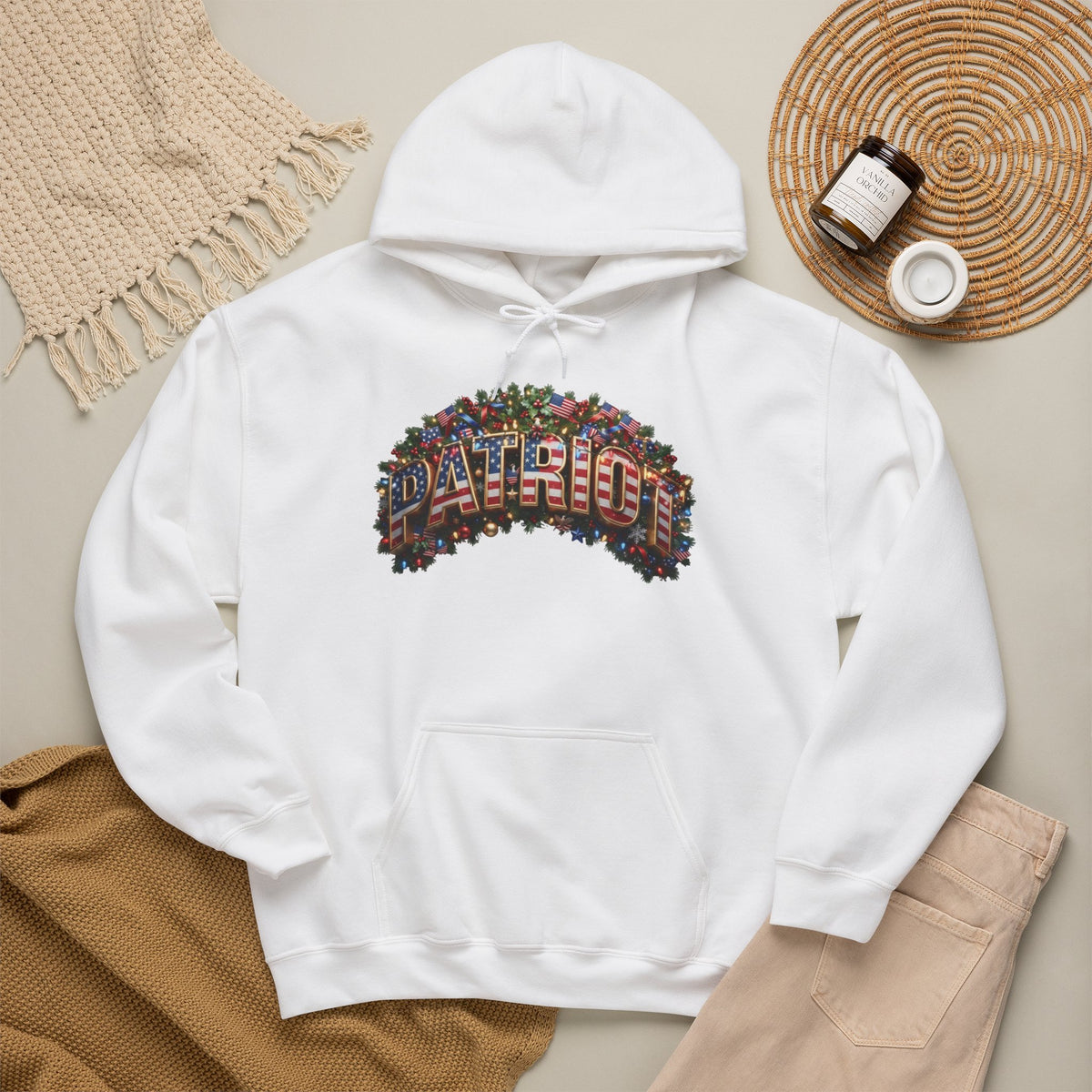 A white pullover hoodie laid flat on a beige surface, featuring a colorful Christmas 'PATRIOT' graphic made from small flag motifs across the chest. The hoodie is styled with a tan knit blanket, beige pants, a woven placemat, and small jars in a flat-lay arrangement.