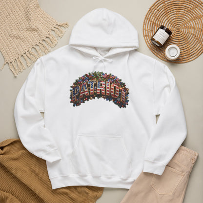A white pullover hoodie laid flat on a beige surface, featuring a colorful Christmas 'PATRIOT' graphic made from small flag motifs across the chest. The hoodie is styled with a tan knit blanket, beige pants, a woven placemat, and small jars in a flat-lay arrangement.