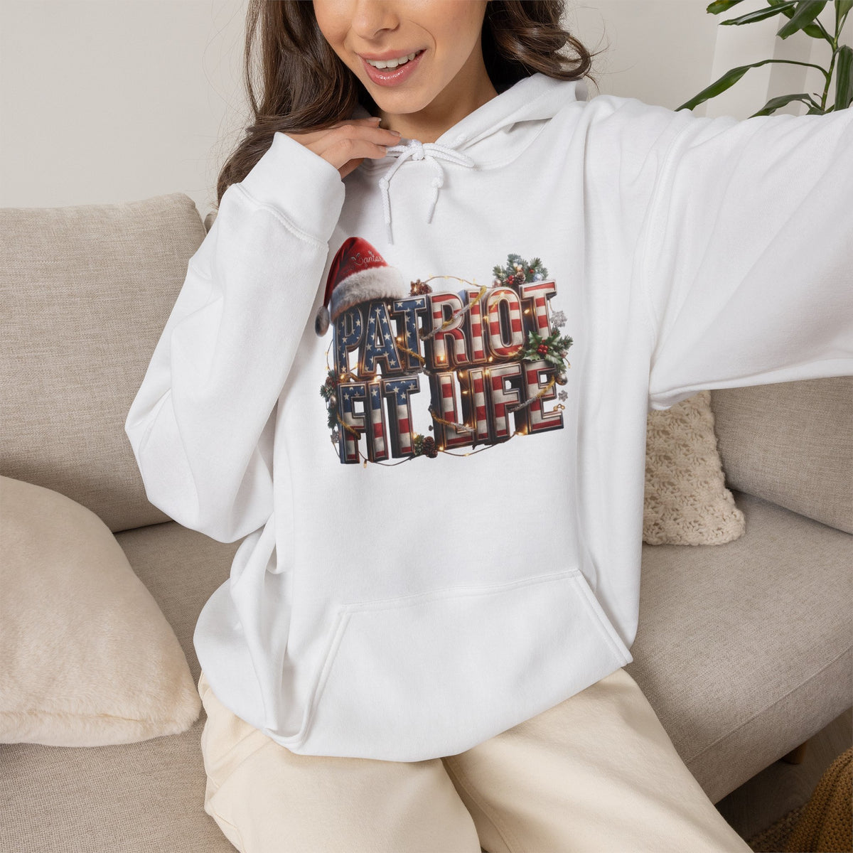 A woman wearing a white pullover hoodie with a large Christmas-themed graphic that reads 'PATRIOT FIT LIFE' in red, green, and gold tones, sitting on a beige couch.