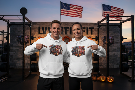 A man and a woman stand in an outdoor gym wearing matching white hoodies with a Halloween graphic that reads 'PATRIOT FIT LIFE. The background features a stone wall with 'LIVE FIT' text, American flags, weight racks, and jack-o'-lanterns.