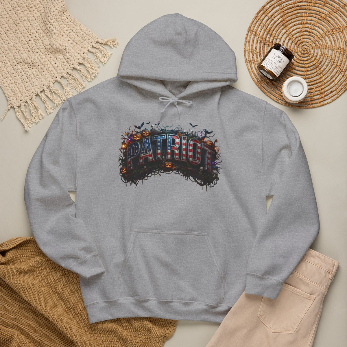 A light gray pullover hoodie with a colorful Halloween 'PATRIOT' graphic design featuring pumpkins, bats, and a distressed texture, styled in a flat lay with a woven blanket, tan sweater, beige pants, and a small candle on a light beige background.