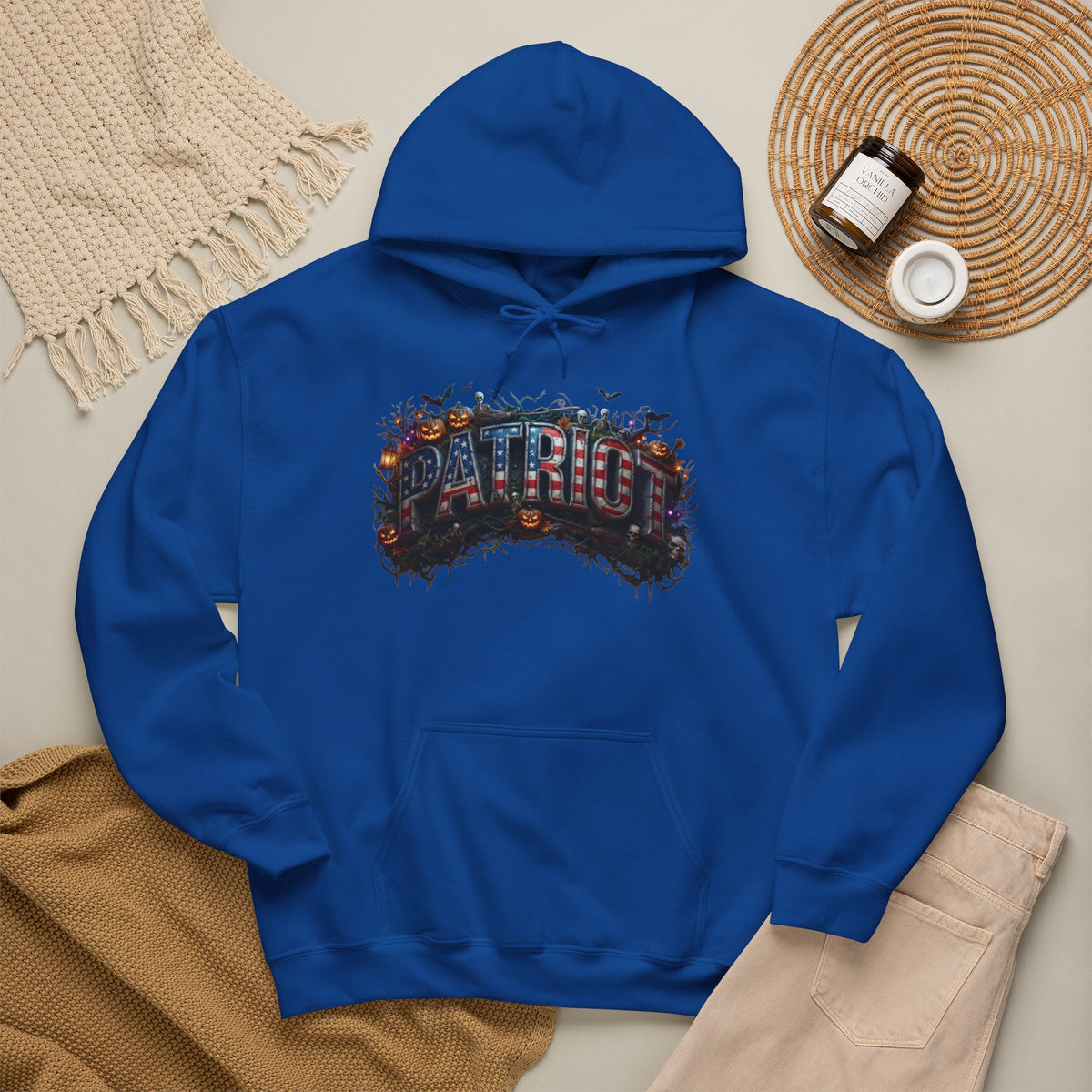 A royal blue hoodie laid flat on a light beige surface, featuring a colorful Halloween 'PATRIOT' graphic design across the chest with autumn and Halloween-themed lettering. The hoodie is styled in a flat lay with a cream knit blanket, tan fabric, beige pants, and a woven round mat with small jars.