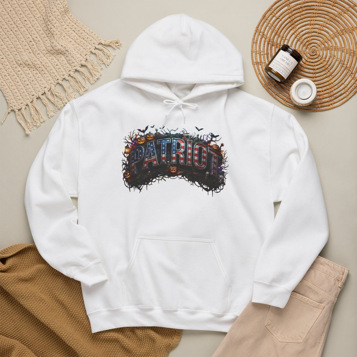 A white pullover hoodie laid flat on a beige surface, featuring a colorful Halloween graphic across the chest with the word “PATRIOT” in stylized letters surrounded by pumpkins, bats, and autumn foliage. The hoodie is styled in a flat lay with a woven blanket, tan pants, a small jar, and a round tray nearby.