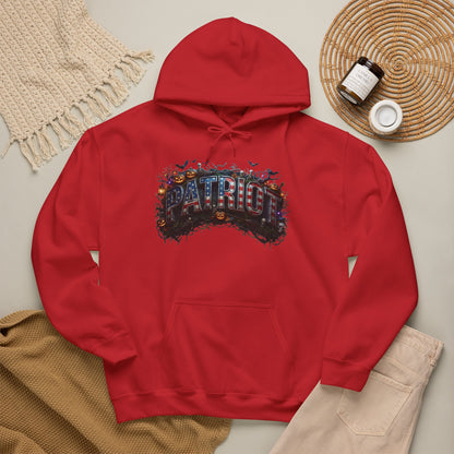 A red pullover hoodie laid flat on a light beige surface, featuring a colorful Halloween 'PATRIOT' graphic design across the chest with a distressed, layered design. The hoodie is styled in a flat lay with a cream knit blanket, a tan knit sweater, beige pants, and a small jar on a woven mat.