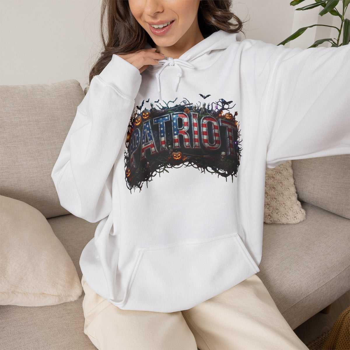 A woman wearing a white hoodie with a large 'PATRIOT' graphic across the chest, featuring a distressed American flag and Halloween theme with pumpkins and bats, sitting on a beige couch.