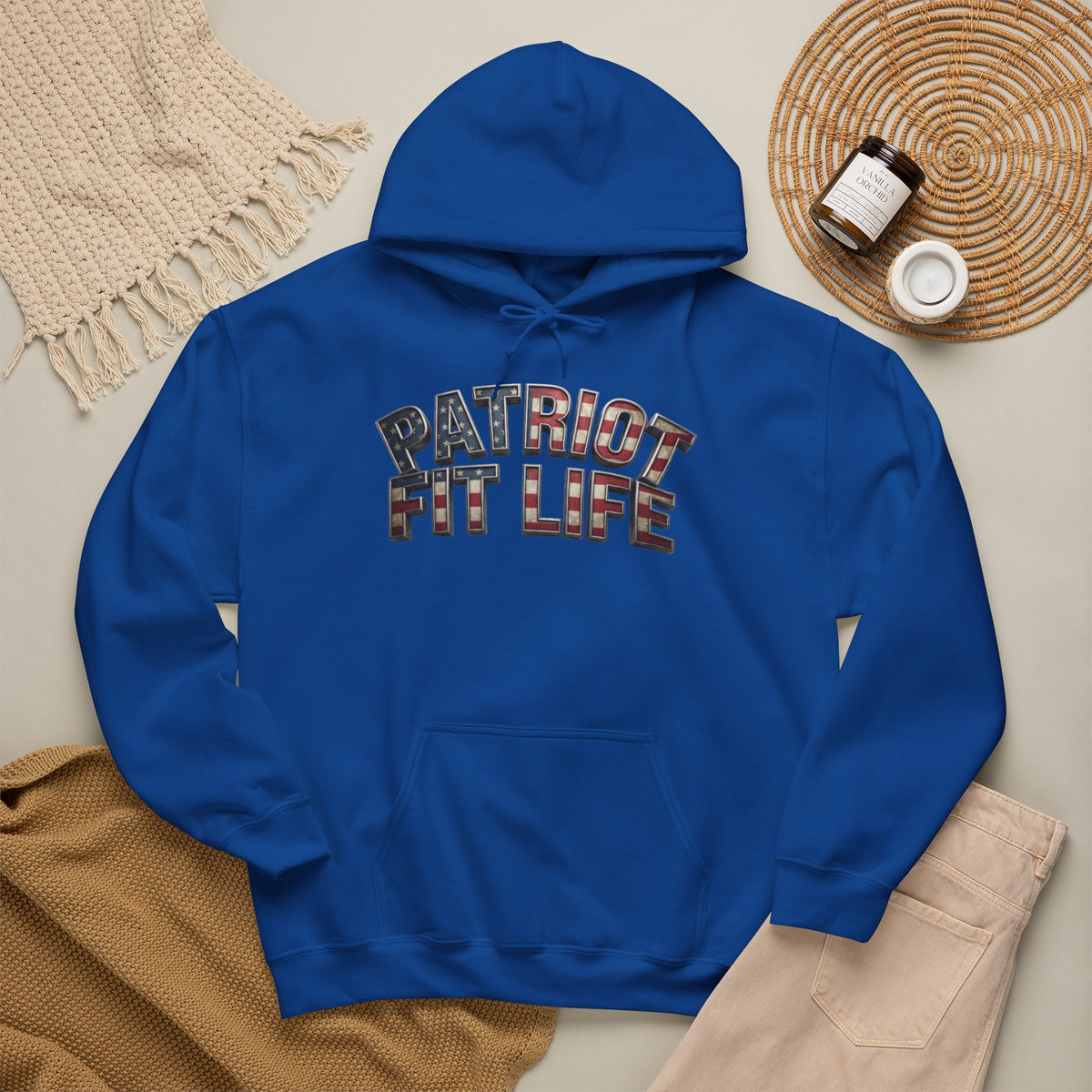 A royal blue pullover hoodie laid flat on a light beige surface, featuring the words 'PATRIOT FIT LIFE' across the chest in a distressed American flag pattern. The hoodie is styled with a cream knit blanket, tan knit fabric, beige pants, and a woven placemat with small accessories.