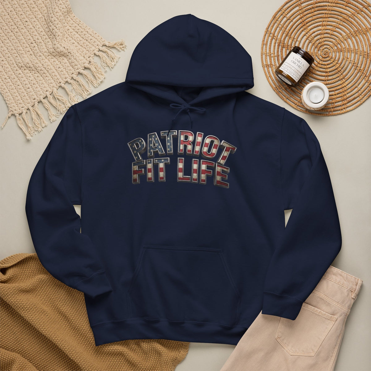 A navy blue pullover hoodie laid flat on a light beige surface, featuring the words 'PATRIOT FIT LIFE' across the chest in a distressed American flag pattern. The hoodie is styled in a flat lay with a cream knit blanket, tan fabric, light pants, and a small jar on a woven mat.