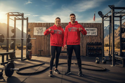 A man and a woman standing in a rustic outdoor gym wearing matching red hoodies with 'PATRIOT FIT LIFE' printed on the front, posing in front of a wooden wall with mountain scenery in the background.