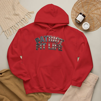 A red pullover hoodie laid flat on a light beige surface, featuring the words 'PATRIOT FIT LIFE' in a plaid pattern across the chest. The hoodie is styled with a cream knit blanket, a tan knit fabric, a woven placemat, and small jars in a cozy flat-lay arrangement.