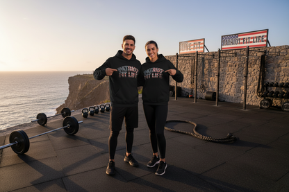 A man and a woman standing on an outdoor cliffside gym floor wearing matching black hoodies with 'PATRIOT FIT LIFE' printed across the chest, pointing at the text. The background shows the ocean, a stone wall, and fitness equipment, including a barbell, kettlebells, battle ropes, and pull-up bars under signs reading 'LIVE FIT' and 'PATRIOT FIT LIFE.'