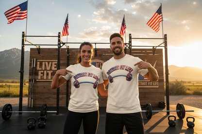 A man and a woman stand outdoors in front of a wooden fitness wall with flags, mountains, and exercise equipment, wearing matching white graphic T-shirts that read 'Patriot Fit Life' with a barbell and patriotic red, white, and blue design.