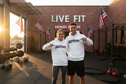 A man and a woman are standing in an outdoor gym wearing matching white hoodies with 'PATRIOT' printed on the front, and they are both pointing at the logos. The background shows a brick wall with 'LIVE FIT' & PATRIOT FIT LIFE signage, American flags, and gym equipment.