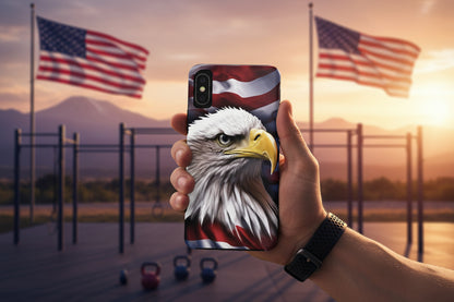 A phone case with a bald eagle American flag design held by a hand, with American flags and a sunset in the background.