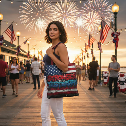 A woman holding a tote bag with text that says, Stronger Than Yesterday and an American flag design on a boardwalk with fireworks in the background.