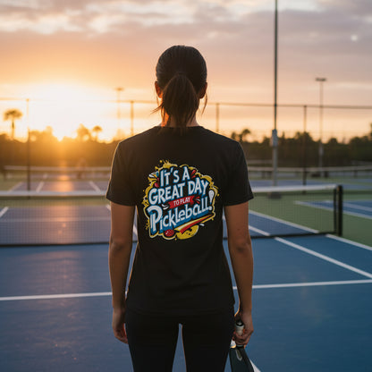 Person on a pickleball court wearing a black 'It's a Great Day to Play Pickleball' shirt at sunset.